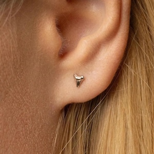 Peut inclure: Boucle d'oreille clou argentée en forme de dent de requin. L'image montre l'oreille et les cheveux blonds. Le bijou est de conception simple et minimaliste, idéal pour un usage quotidien.