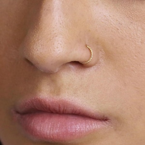 May include: Close-up of a gold nose ring. The ring is a small, twisted hoop, and is worn on the right nostril. The skin around the nose and mouth is visible.
