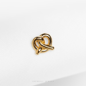 May include: A gold-coloured pretzel charm. The charm is a miniature representation of a pretzel, with its characteristic looped shape. The pretzel is made of a shiny, metallic material, and is set against a white background.