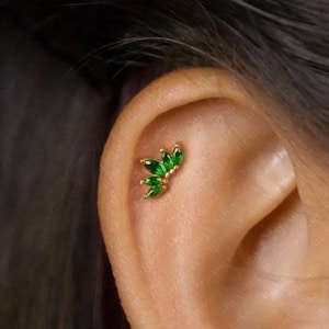 Peut inclure: Un piercing d'oreille doré orné d'un groupe de pierres précieuses vertes taillées en émeraude. Les pierres sont disposées en forme de fleur ou de feuille, ajoutant une touche d'élégance au bijou.