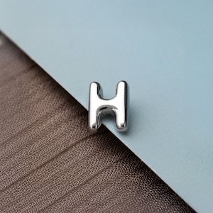 May include: A polished silver-toned metal charm shaped like the capital letter H. The letter features a smooth, rounded, and reflective surface with a three-dimensional block design. The piece is solid in colour and has a clean, uniform metallic finish.