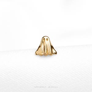 May include: A small gold-toned stud earring shaped like a ghost with two circular cutouts for eyes and a scalloped bottom edge. The piece features a polished metallic finish and is positioned on a white fabric surface with the text SHEMISET JEWELS.