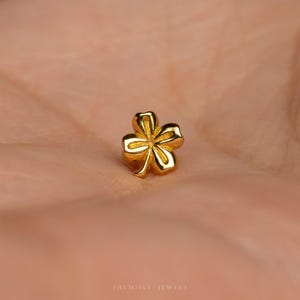 May include: A small, polished gold stud earring shaped like a four-leaf clover. The piece features a smooth, reflective metallic surface with defined indentations separating each of the four rounded petals, creating a classic, symmetrical floral design.