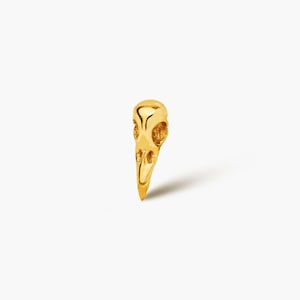 May include: A gold-coloured raven skull pendant. The pendant is highly detailed, with visible eye sockets and a pointed beak. The skull is cast in a shiny, metallic finish, and is set against a white background.