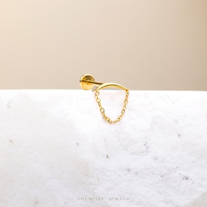 May include: A gold labret stud featuring a curved bar and a delicate chain. The jewellery is presented on a white surface against a neutral backdrop. The brand name "SHEMISLI JEWELS" is printed at the bottom.