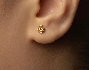 May include: A close-up of a gold spiral stud earring on an ear. The earring is small and circular, with a coiled design. The earring is a bright gold colour and appears to be made of metal. The text "SHEMISLI JEWELS" is at the bottom of the image.
