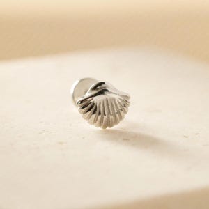 May include: A silver seashell stud earring. The earring has a detailed, textured seashell design. The stud post is visible on the back. The earring is set against a neutral background.