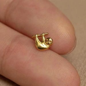 May include: A tiny, gold-coloured sloth charm suspended from a horizontal bar. The sloth has a rounded body and a small head. The charm is likely made of metal. The charm is small, with visible details.