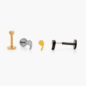 May include: Four pieces of body jewellery are displayed. They include a gold-coloured labret stud, a silver-coloured stud with a comma design, a gold-coloured comma stud, and a black barbell stud.