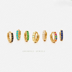 Peut inclure: Une collection de créoles dorées, chacune ornée d'une rangée de pierres précieuses colorées. Les boucles d'oreilles présentent des pierres noires, turquoise, vertes, crème, blanches, arc-en-ciel et bleues. La marque "SHEMISLI JEWELS" est imprimée en dessous.