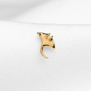 May include: A small, gold-coloured stingray-shaped pin. The pin has a curved tail and detailed fins. The pin is set against a white background, highlighting its metallic sheen. This jewellery item is suitable for adding a touch of marine-inspired flair to any outfit.