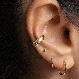 Peut inclure: Gros plan d'une oreille ornée de bijoux en or. L'oreille présente un ear cuff en or, un petit clou d'oreille en or et deux créoles en or. Les bijoux ont une surface polie et réfléchissante.