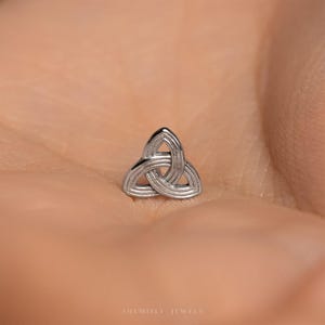 May include: A small, silver Triquetra symbol, a Celtic knot design, is shown. The three-lobed knot is a continuous line, representing interconnectedness. The jewellery piece is set against a neutral background.
