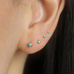 May include: Four gold stud earrings with turquoise stones of different sizes are shown on an ear. The earrings have a gold setting and the turquoise stones are a light blue colour. The earrings are of varying sizes.