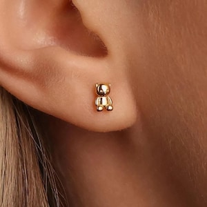 May include: A gold teddy bear stud earring. The earring is small and detailed, shaped like a bear. The earring is worn on an ear. The background is blurred, focusing attention on the earring.