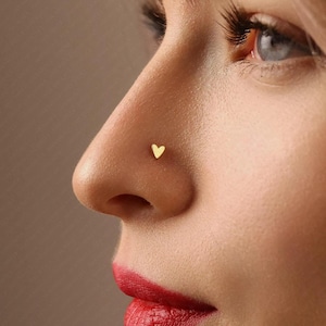 Peut inclure: Gros plan sur un nez percé d'un petit bijou en forme de cœur doré. La peau est claire et les lèvres sont peintes d'un rouge vif. L'arrière-plan est d'une teinte neutre et douce.