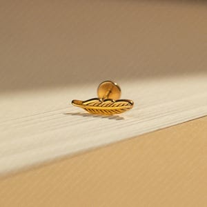 May include: A gold-coloured feather-shaped earring stud. The earring has a post backing and is set against a neutral background. The feather design features detailed lines, giving it a realistic appearance. This jewellery piece is suitable for everyday wear.