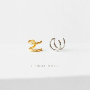 May include: Two ear cuffs, one gold and one silver, are shown against a white backdrop. The gold cuff has a spiral design, while the silver cuff has a three-loop design. The text "SHEMISLI JEWELS" is at the bottom of the image.
