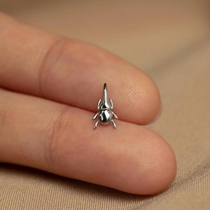 Peut inclure: Une petite boucle d'oreille argentée en forme de scarabée. La boucle d'oreille est très détaillée, avec des pattes, des antennes et un corps texturé visibles. Le scarabée est posé sur un doigt, mettant en valeur sa taille et son design. Le fond est beige neutre.