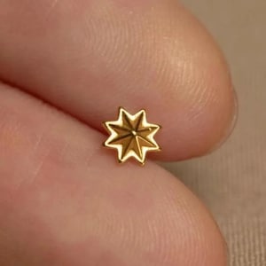 May include: A small, gold-coloured star-shaped stud earring. The earring has eight points and a slightly textured surface. The star is centered and appears to be made of metal.