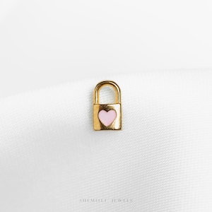 Peut inclure: Un petit charm en forme de cadenas doré avec un cœur rose au centre. Le charm est posé sur un fond blanc texturé. Le cadenas a une anse arrondie et un corps rectangulaire. Probablement pour la bijouterie.