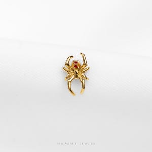 May include: A gold-coloured spider-shaped jewellery piece with a red accent on its back. The spider has eight legs and is set against a white background. The words "SHEMISLI JEWELS" are at the bottom.