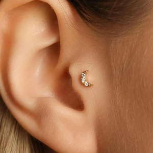 May include: A gold-coloured crescent moon earring with three clear, round gemstones. The earring is small and delicate, designed for a cartilage piercing. The earring is shown on a person's ear.