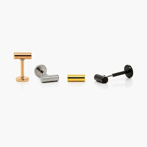 May include: Four lip piercing studs in gold, silver, yellow gold, and black. The studs have a flat back and a cylindrical front. The gold stud is upright, while the others are lying down.