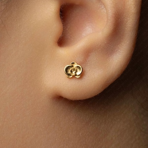 Peut inclure: Une petite boucle d'oreille en forme de fleur de couleur or est montrée dans l'oreille. La boucle d'oreille a un design détaillé avec de multiples pétales et un centre texturé. C'est un bijou simple et élégant.
