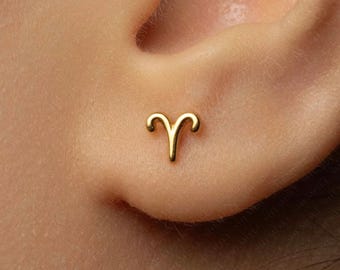 Könnte beinhalten: Ein kleiner Ohrstecker aus poliertem Gold in Form des Tierkreiszeichens Widder. Das Schmuckstück hat eine glatte, metallische Oberfläche und ist so gestaltet, dass es flach am Ohrläppchen anliegt und die geschwungenen Hörner zeigt.