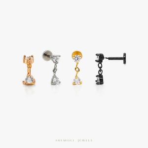 May include: Four stud earrings in gold, silver, and black, each with a dangling design and a clear, sparkling stone. The earrings are displayed against a white backdrop. The brand name "SHEMISLI JEWELS" is visible at the bottom of the image.