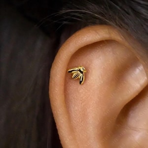 Peut inclure: Une boucle d'oreille dorée en forme d'abeille. La boucle d'oreille est petite et détaillée, avec des accents noirs sur les ailes et le corps. La boucle d'oreille est montrée sur une oreille, soulignant sa taille et son design.