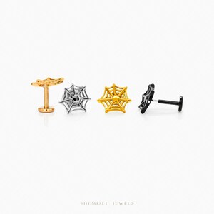 May include: Four spiderweb-shaped earrings in gold, silver, and black. The earrings are displayed on a white background. The earrings are small and delicate, with a detailed web design. The brand name "SHEMISLI JEWELS" is at the bottom.