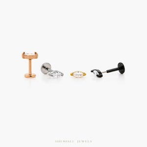 May include: Four pieces of jewellery are displayed against a white background. The jewellery includes a rose gold stud with a curved top, a silver stud, a gold stud, and a black stud. Each stud features a clear, marquise-cut gemstone.
