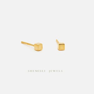May include: Two gold cube stud earrings on a white backdrop. The earrings are small and minimalist, with a simple geometric design. The text "SHEMISLI JEWELS" is printed below the earrings.