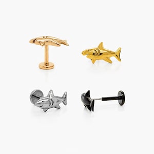 May include: Four shark-themed body jewellery pieces in gold, silver, and black. The jewellery includes a gold shark stud, a gold shark with a post, a silver shark stud, and a black shark with a post.
