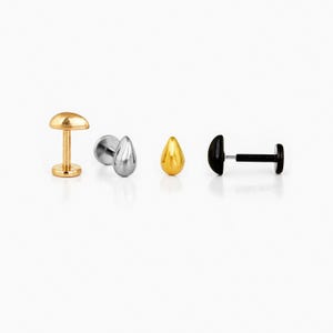 May include: Four pieces of body jewellery are displayed against a white background. The pieces include a gold-coloured stud, a silver teardrop-shaped stud, a gold teardrop-shaped stud, and a black stud with a post.