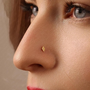 Peut inclure: Gros plan sur un nez avec un petit piercing de nez en forme de losange de couleur or. La peau est claire avec des pores visibles et une légère teinte rosée. L'accent est mis sur le nez et le bijou.
