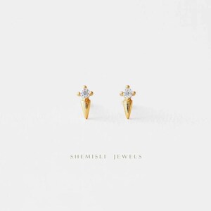 May include: A pair of gold stud earrings. Each earring has a conical base and a small, round, clear gemstone. The earrings are set against a white backdrop. The text "SHEMISLI JEWELS" is printed below the earrings.