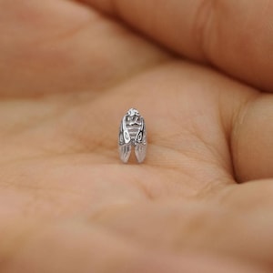 May include: A small, silver charm with an insect design. The charm features detailed wings and a textured body, resembling a cicada or similar insect. The charm is set against a blurred background.
