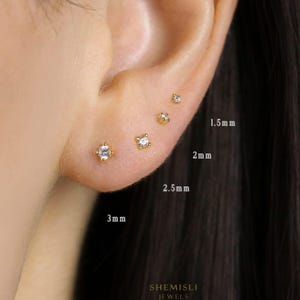 May include: An ear adorned with four gold stud earrings, each with a square, clear gemstone. The earrings are of varying sizes, labelled as 1.5mm, 2mm, 2.5mm, and 3mm. The brand name "SHEMISLI JEWELS" is visible at the bottom of the image.