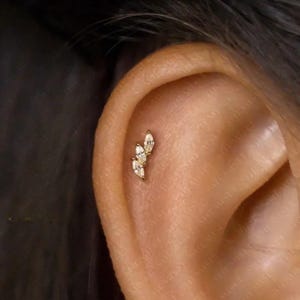 May include: A gold-coloured ear piercing featuring three marquise-cut, clear gemstones. The piercing is arranged in a cluster design, offering a delicate and elegant appearance. The jewellery is shown on an ear.