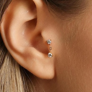 May include: A gold earring featuring two clear, round gemstones linked by a fine chain. The image provides a close-up view, emphasizing the sparkle of the stones and the intricate design of the jewellery.