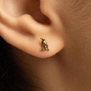 May include: A small, gold-coloured stud earring shaped like a bird, specifically a raven or crow, featuring a polished metallic finish. The earring is positioned on the earlobe, showcasing the detailed silhouette of the bird in a standing pose.