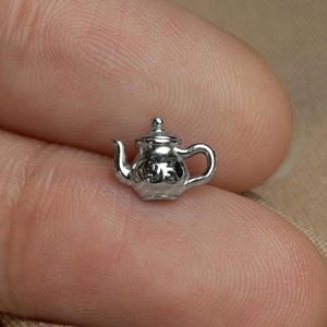 May include: A miniature silver teapot charm with intricate detailing. The teapot features a curved spout, a handle, and a lid with a small knob. The charm is likely made of metal and is set against a neutral background.