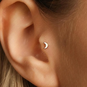 May include: A small, gold-coloured crescent moon stud earring. The earring has a minimalist design with a smooth, polished surface. The earring is shown on an ear.