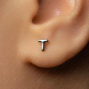 May include: A small, polished silver-toned stud earring shaped like the capital letter T. The earring features a smooth, rounded surface and is positioned securely against the skin of an earlobe, showcasing a simple and clean metallic design.