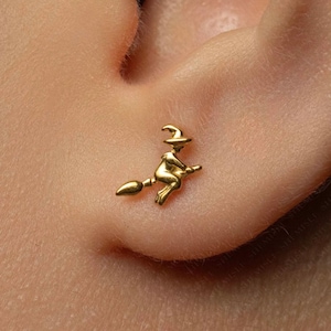 Peut inclure: Une petite boucle d'oreille dorée en forme de sorcière portant un chapeau pointu et chevauchant un balai. La silhouette est représentée de profil, mettant en évidence la forme du chapeau, le corps de la sorcière et le manche du balai.