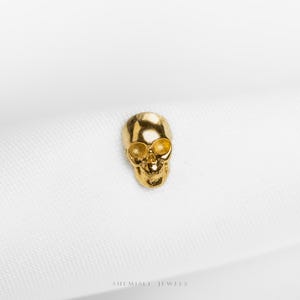 May include: A small, gold-coloured skull charm with detailed facial features. The skull has eye sockets and a nose cavity. The charm is set against a white background. The jewellery piece is likely a pendant or earring.