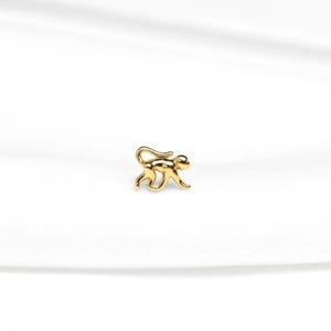 May include: A small, gold-coloured monkey charm. The charm is a detailed, three-dimensional rendering of a monkey in a walking pose. The monkey's tail is curled, and its limbs are extended. The charm is set against a white background.